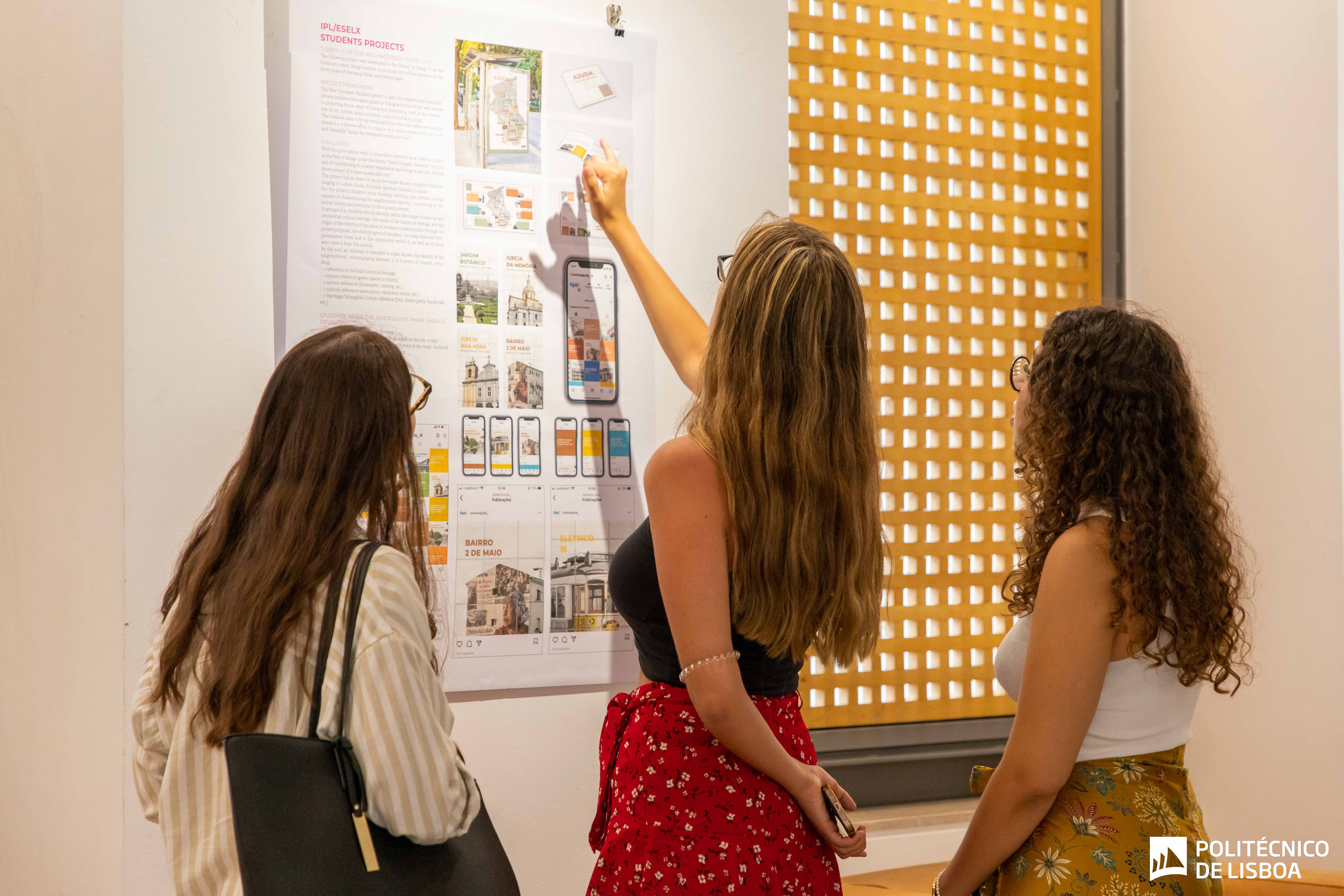 foto de 3 raparigas de pé a observar um painel pendurado na parede com trabalhos de designs