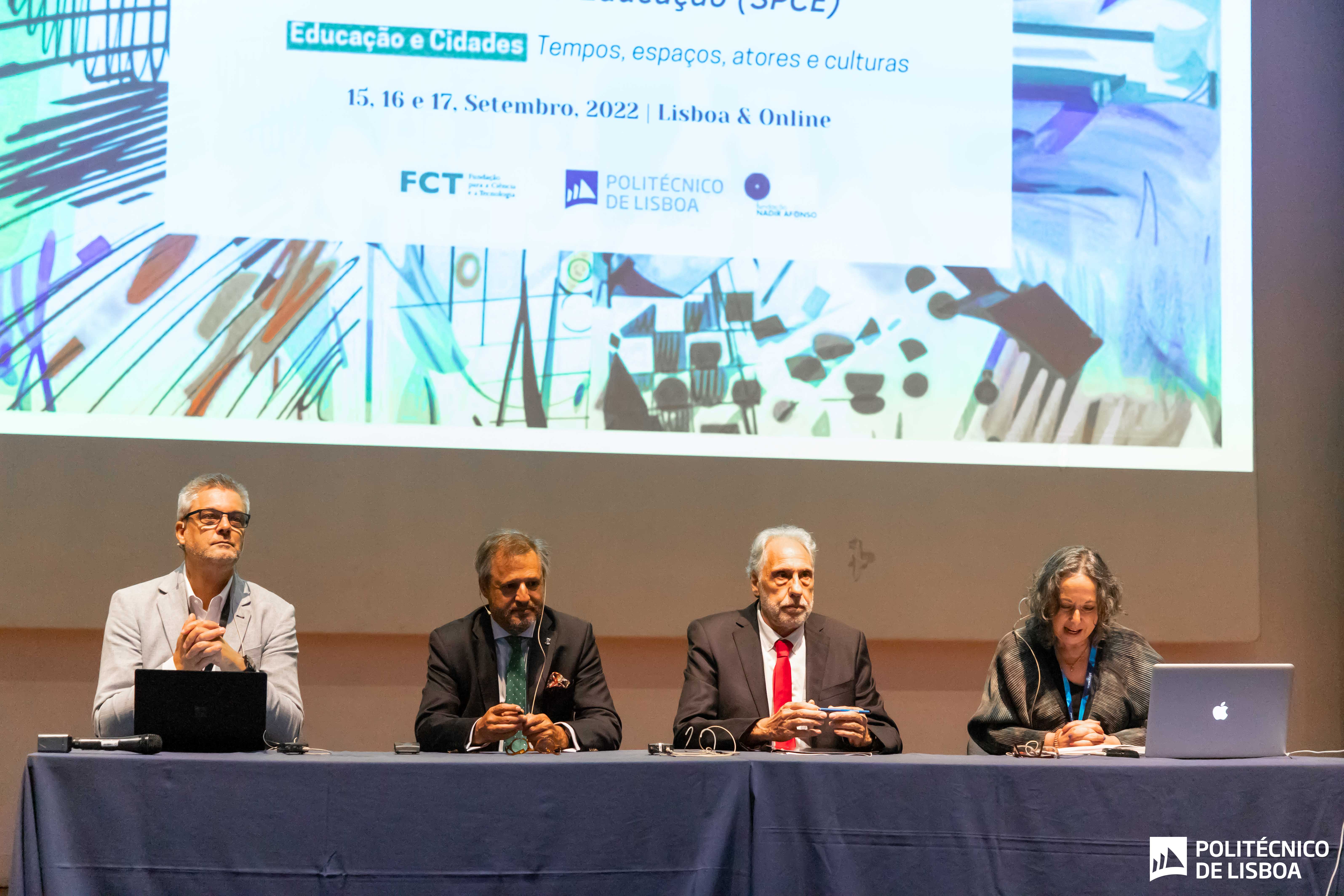 3 homens e 1 mulher sentados numa mesa num palco com uma projeção