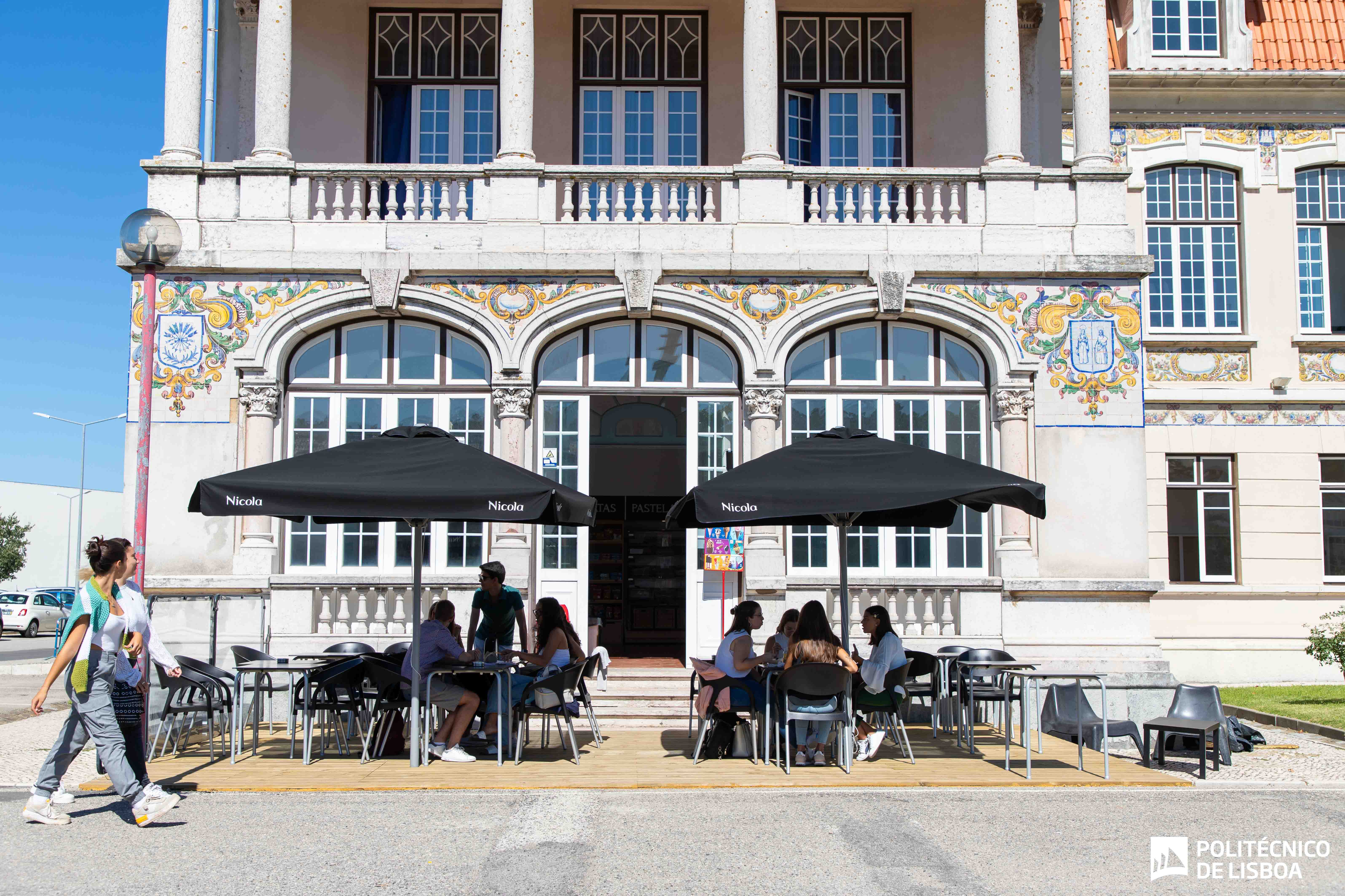 esplanada de bar com chapéus e pessoas sentadas nas mesas e pessoas a passar a pé