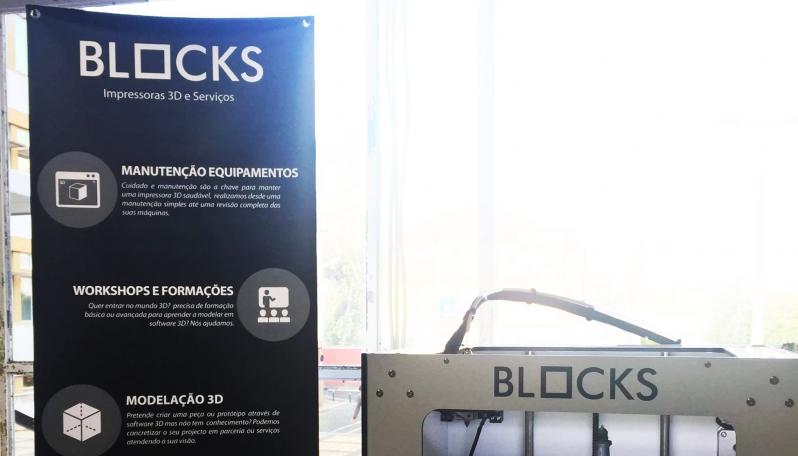 Imagem de um roll-up mencionando a marca Blocks ao lado de uma impressora 3D