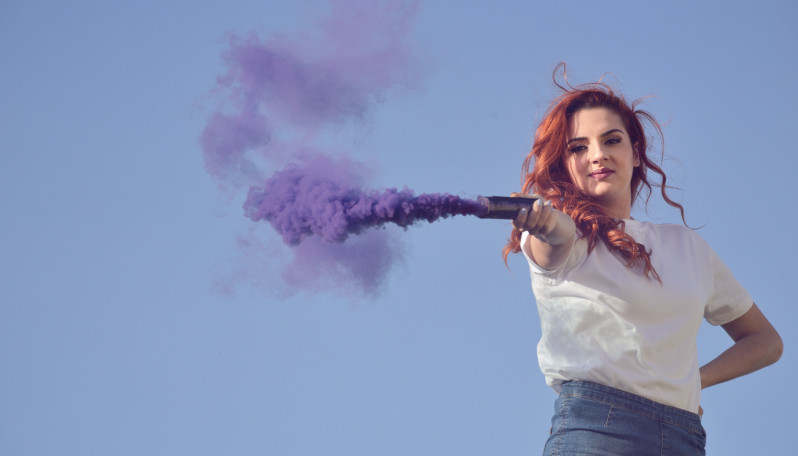 Imagem onde se vê uma rapariga de cabelo vermelho e de t-shirt branca a segurar uma tocha de fumo roxo