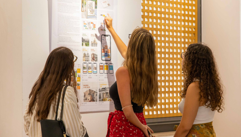 foto de 3 raparigas de pé a observar um painel pendurado na parede com trabalhos de designs 