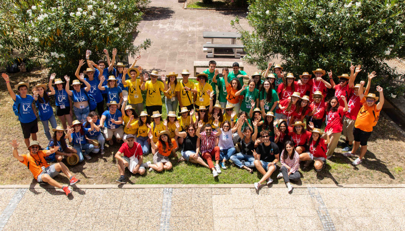 foto com dezenas de jovens com t-shirts de várias cores em pé e sentados num jardim de baços no ar