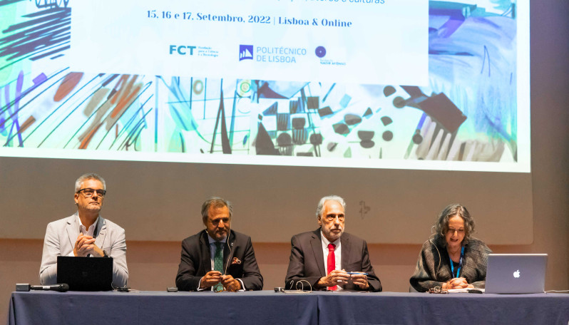 3 homens e 1 mulher sentados numa mesa num palco com uma projeção