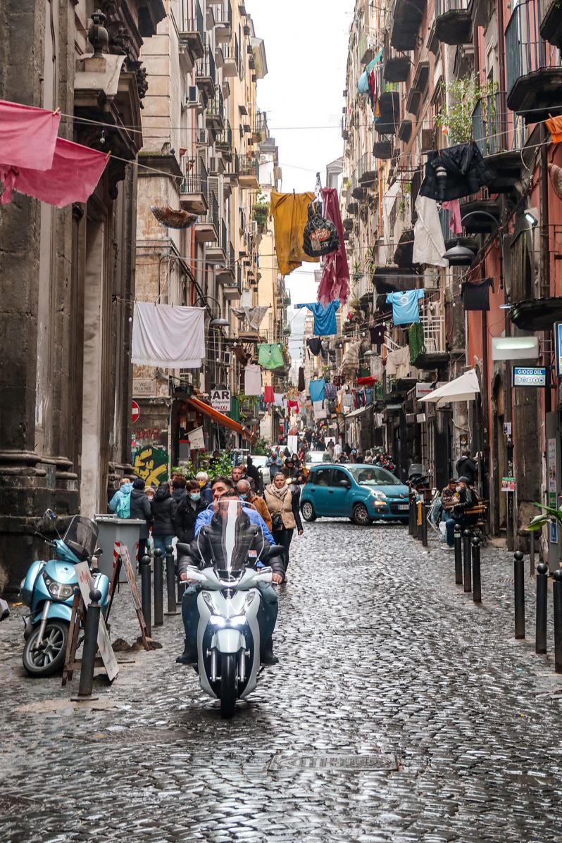 rua de cidade italiana com vespa a andar e roupa colorida pendurada nas casas