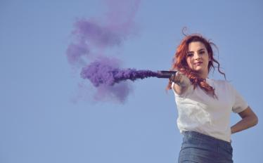 Imagem onde se vê uma rapariga de cabelo vermelho e de t-shirt branca a segurar uma tocha de fumo roxo