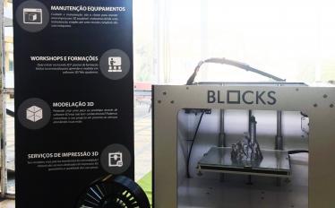 Imagem de um roll-up mencionando a marca Blocks ao lado de uma impressora 3D