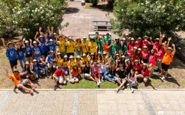 foto com dezenas de jovens com t-shirts de várias cores em pé e sentados num jardim de baços no ar